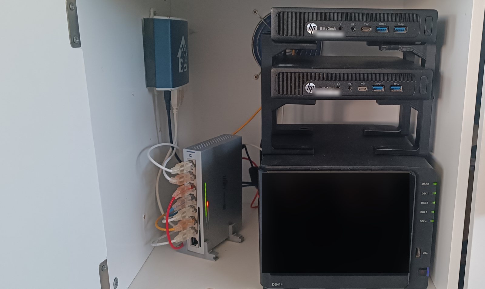 Two HP EliteDesk Mini nodes on stacking stands above a Synology DS414 NAS, with a UniFi switch and the Home Assistant box beside them in a white cupboard